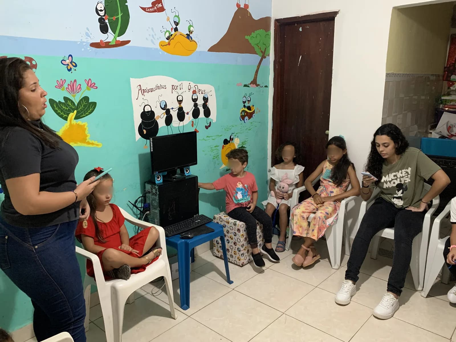 Crianças sentadas em roda numa sala de paredes pintadas, ouvindo a professora da Classe Kids
