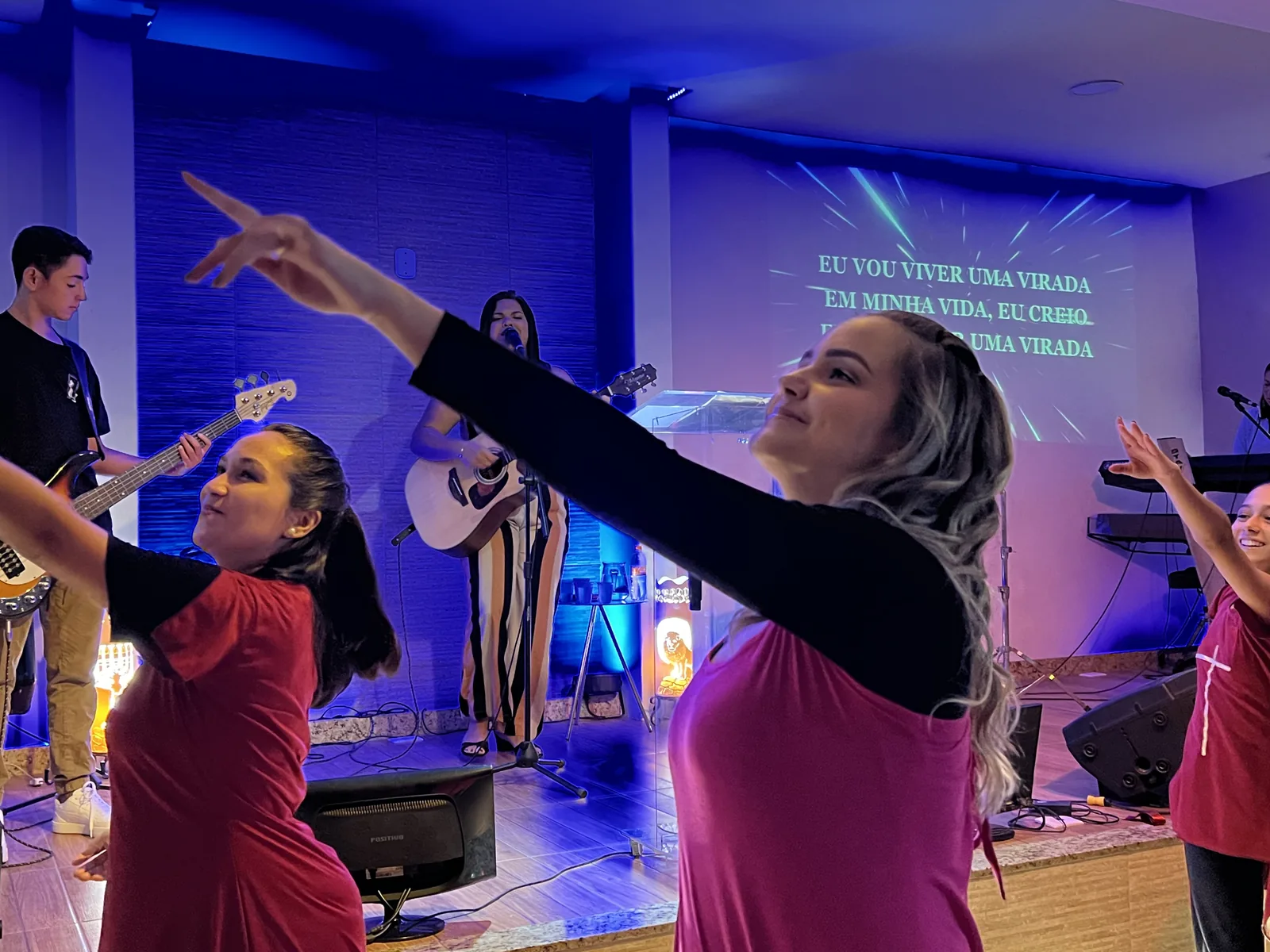 Mulheres dançando com os braços estendidos no louvor, com a banda e a letra da música ao fundo
